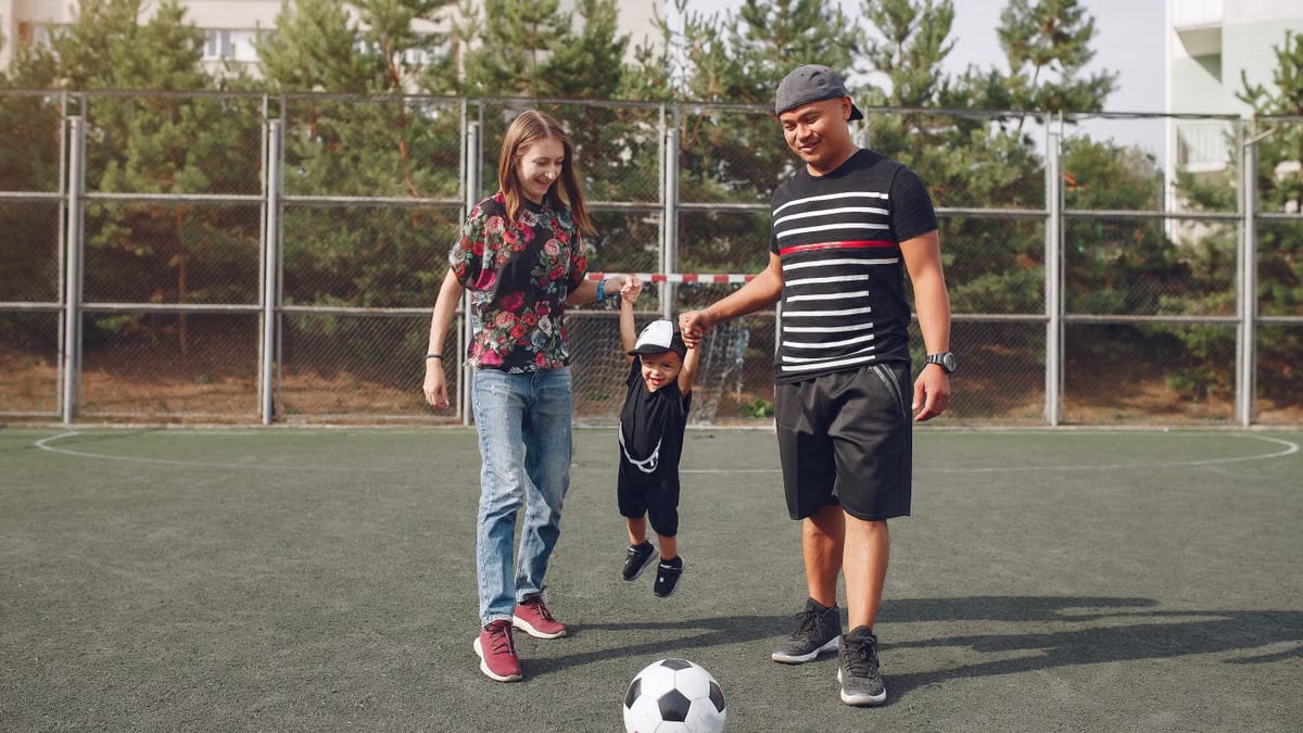 Full body of positive content diverse parents holding son hands and teaching boy to kick ball on football field on sunny summer day